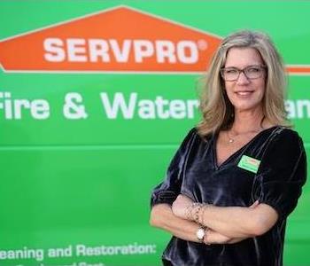 Middle-aged woman with blond, shoulder-length hair, glasses, and blue eyes, wearing a velour, v-neck shirt by a SERVPRO truck